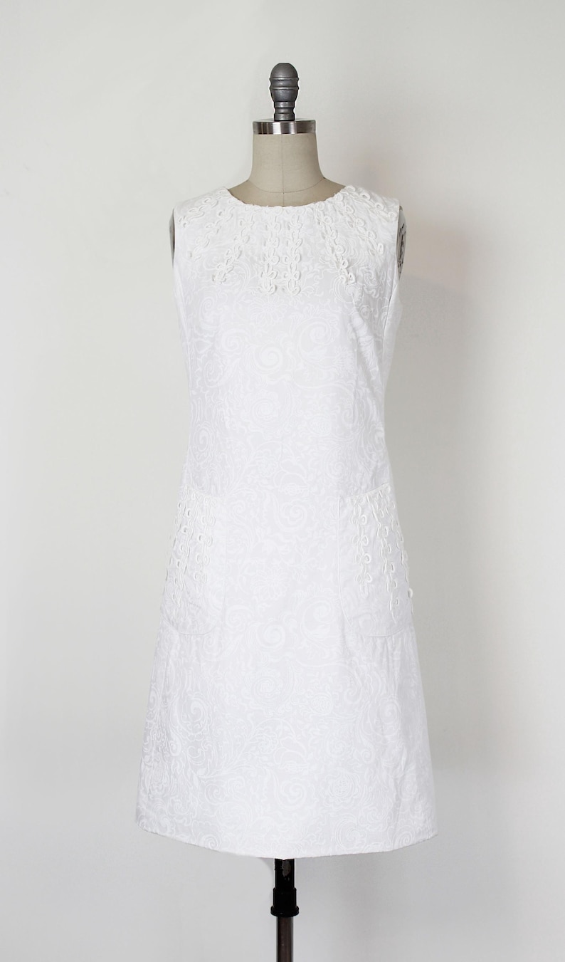 May include: A sleeveless white dress with an intricate floral pattern and eyelet detailing. The dress is displayed on a mannequin, showcasing its A-line silhouette and round neckline. The fabric appears to be cotton or a similar lightweight material.