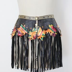 May include: Black leather belt with colorful geometric accents and long fringe. The belt features a decorative gold-toned buckle and is adorned with black beads. The colorful accents include orange, pink, green, and yellow.