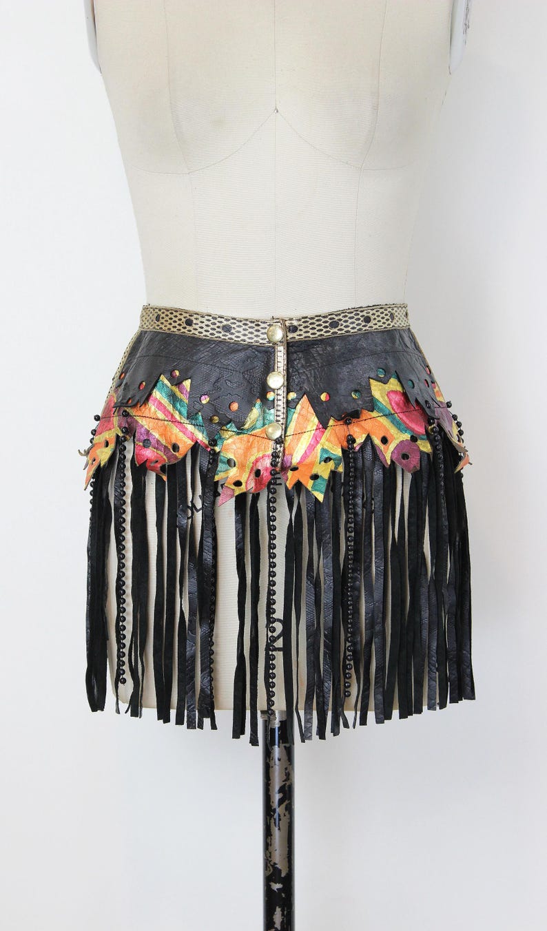 May include: Black leather belt with colorful geometric accents and long fringe. The belt features a decorative gold-toned buckle and is adorned with black beads. The colorful accents include orange, pink, green, and yellow.