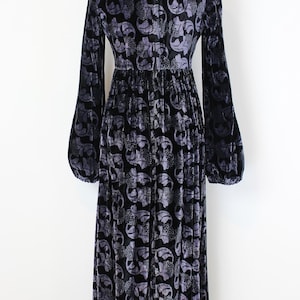 May include: A long, black velvet dress with a floral pattern in shades of gray. The dress features long sleeves with a slight bell shape at the wrist and a fitted waist. The dress is displayed on a mannequin.