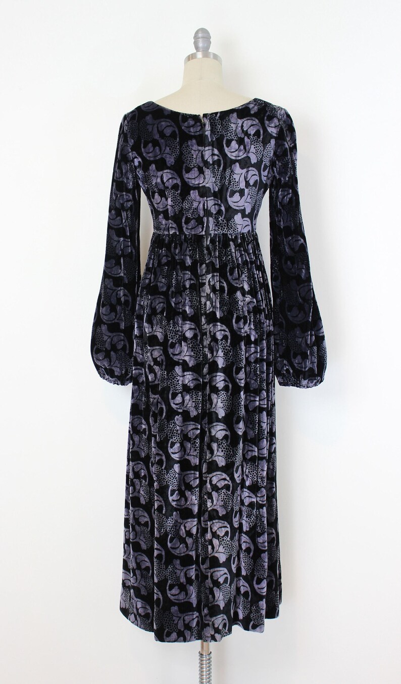 May include: A long, black velvet dress with a floral pattern in shades of gray. The dress features long sleeves with a slight bell shape at the wrist and a fitted waist. The dress is displayed on a mannequin.