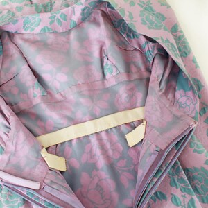 May include: Close-up of a vintage garment with a floral pattern. The fabric features large pink roses with teal green leaves on a lavender background. The lining is a gray fabric with pink roses.