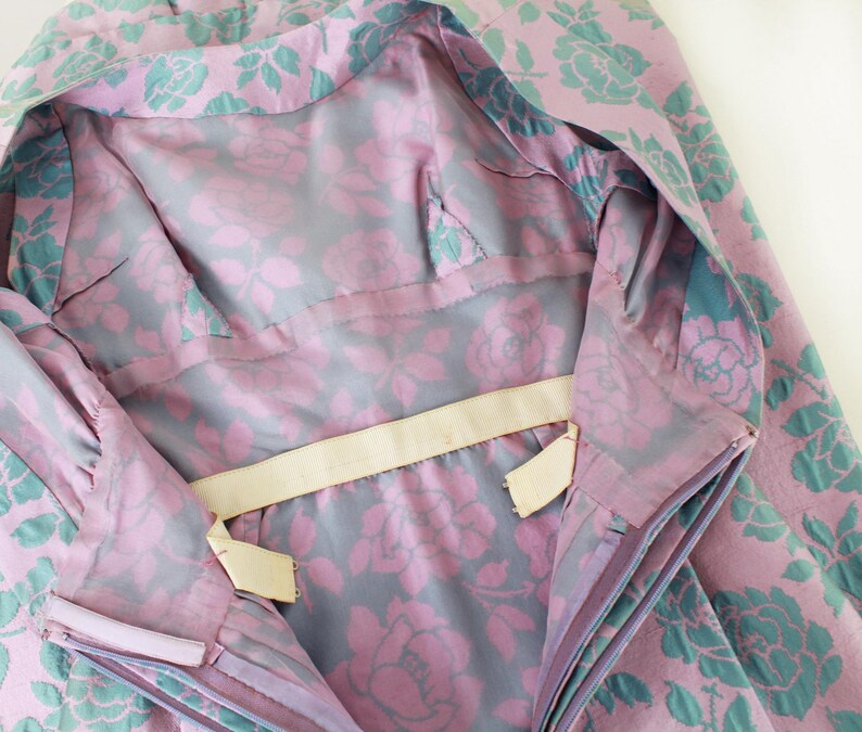May include: Close-up of a vintage garment with a floral pattern. The fabric features large pink roses with teal green leaves on a lavender background. The lining is a gray fabric with pink roses.