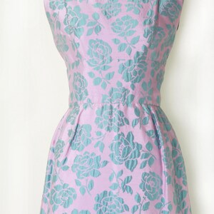 May include: A sleeveless, vintage-style dress in a light pink fabric, adorned with a repeating pattern of teal-colored roses. The dress features a fitted bodice, a defined waist, and a flared skirt, showcasing a classic silhouette.