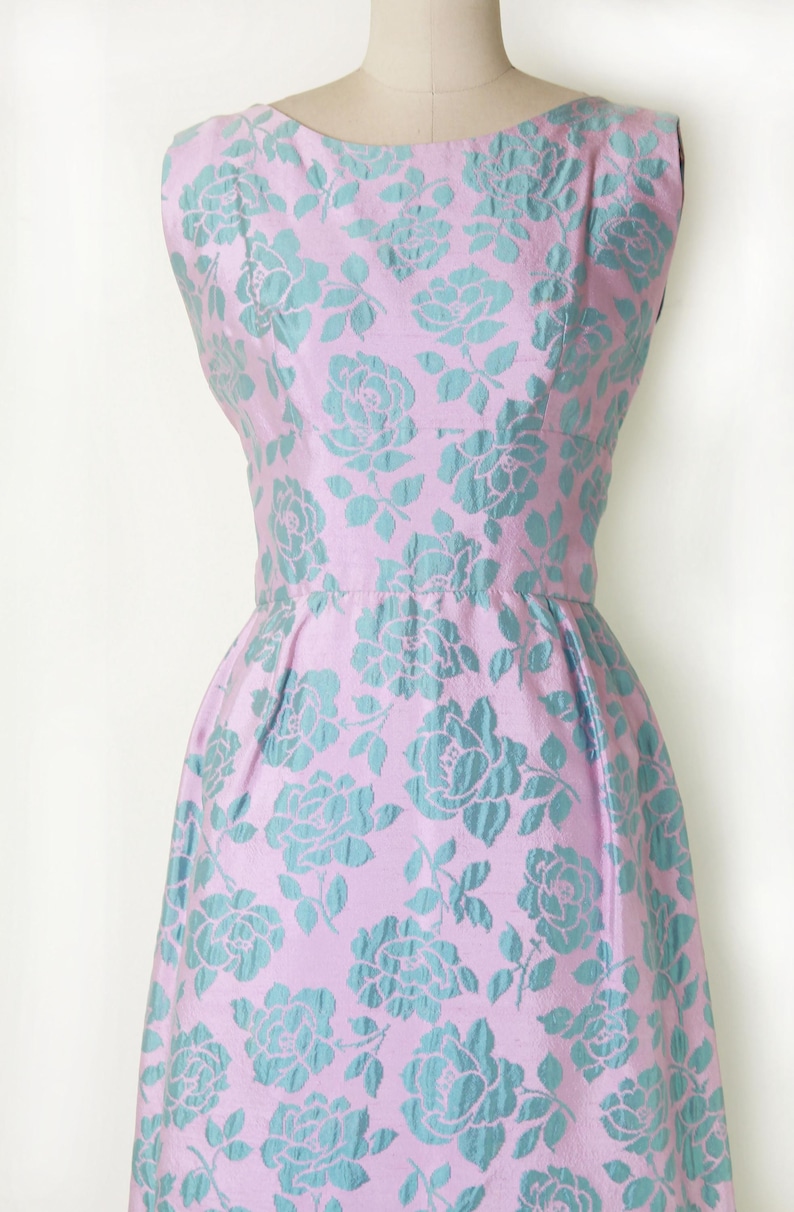 May include: A sleeveless, vintage-style dress in a light pink fabric, adorned with a repeating pattern of teal-colored roses. The dress features a fitted bodice, a defined waist, and a flared skirt, showcasing a classic silhouette.