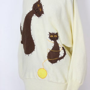 May include: A cream-colored knitted sweater with brown and yellow embroidered cats and a yellow yarn ball.