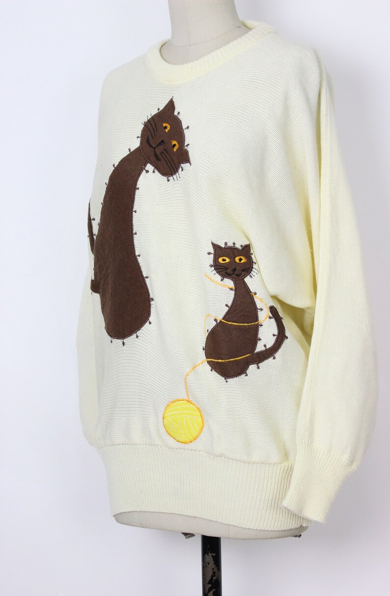 May include: A cream-colored knitted sweater with brown and yellow embroidered cats and a yellow yarn ball.