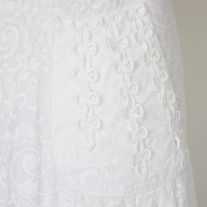 May include: White fabric with a subtle floral pattern. A pocket is adorned with a delicate, white, embroidered design. The fabric appears to be cotton or a similar material, suitable for clothing.
