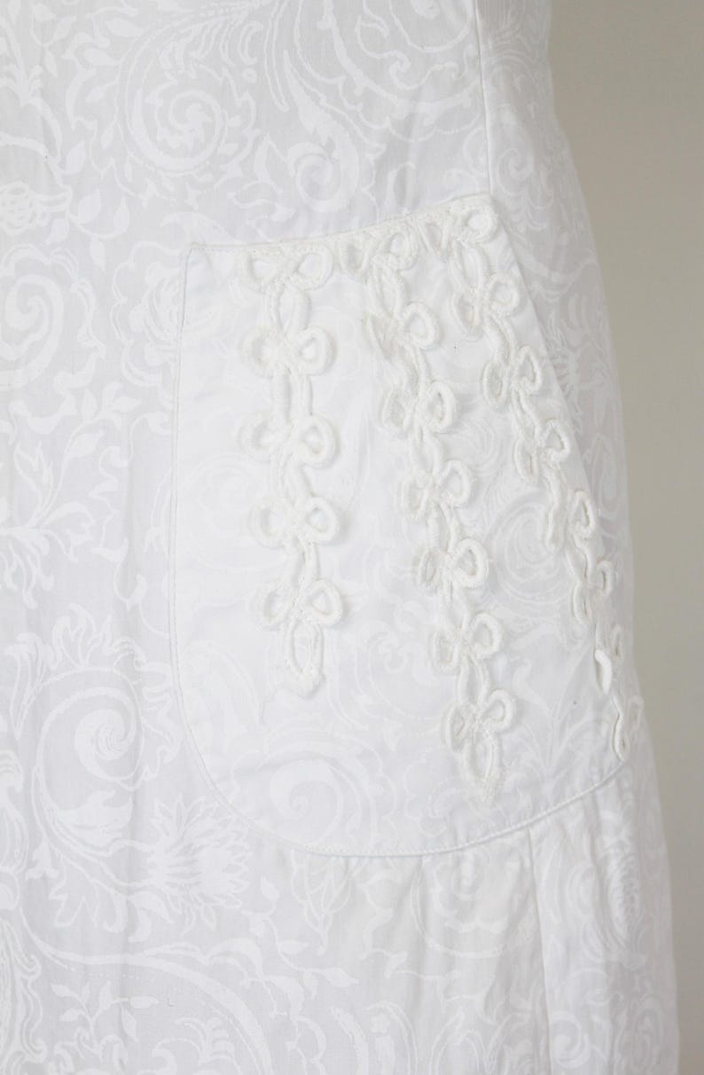 May include: White fabric with a subtle floral pattern. A pocket is adorned with a delicate, white, embroidered design. The fabric appears to be cotton or a similar material, suitable for clothing.