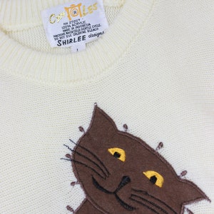 May include: A white knitted sweater with a brown cat embroidered on the front. The sweater has a tag that reads "COLES RN 27267 100% ACRYLIC MADE IN INDONESIA MACHINE WASH COLD GENTLE CYCLE DO NOT USE CHLORINE BLEACH SHIRLEE designs".