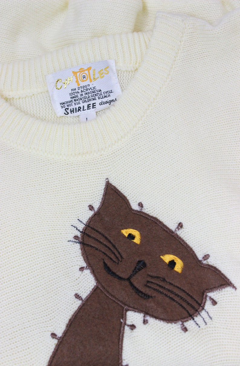 May include: A white knitted sweater with a brown cat embroidered on the front. The sweater has a tag that reads "COLES RN 27267 100% ACRYLIC MADE IN INDONESIA MACHINE WASH COLD GENTLE CYCLE DO NOT USE CHLORINE BLEACH SHIRLEE designs".