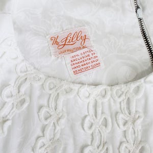 May include: White dress with a floral pattern and decorative openwork trim. The dress features a zipper and a tag that reads "The Lilly" and "100% Cotton Exclusive of Ornamentation Hand Wash Only Do Not Dry Clean."