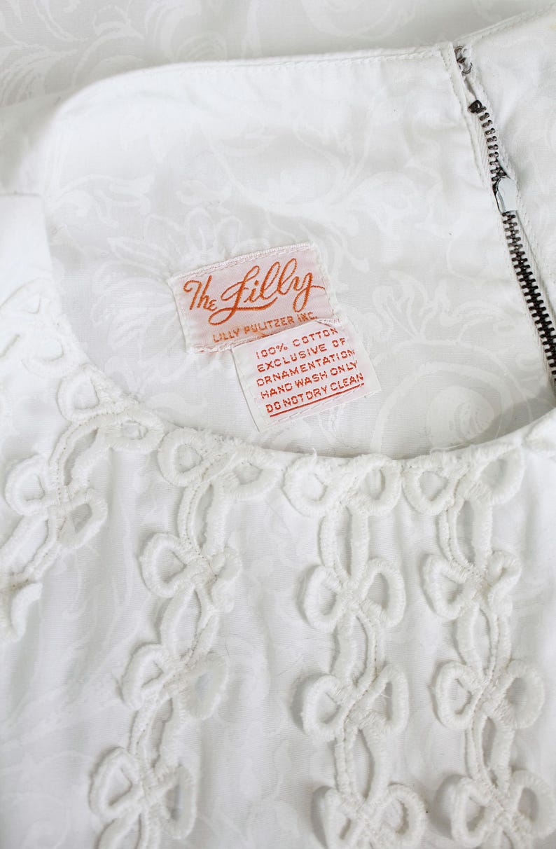 May include: White dress with a floral pattern and decorative openwork trim. The dress features a zipper and a tag that reads "The Lilly" and "100% Cotton Exclusive of Ornamentation Hand Wash Only Do Not Dry Clean."