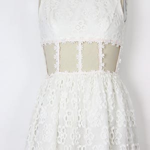 May include: A white lace dress with a high neckline and sleeveless design. The dress has a fitted bodice with a sheer panel at the waist, embellished with floral lace. The skirt flows from the waist, creating a classic shape. The dress is displayed on a mannequin.