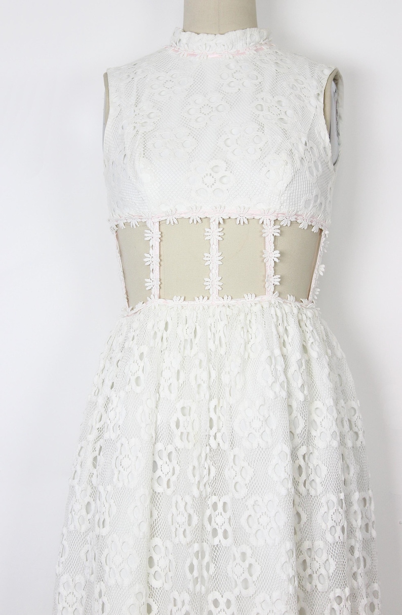 May include: A white lace dress with a high neckline and sleeveless design. The dress has a fitted bodice with a sheer panel at the waist, embellished with floral lace. The skirt flows from the waist, creating a classic shape. The dress is displayed on a mannequin.