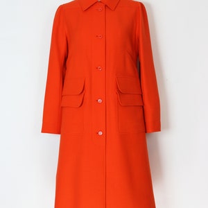 May include: A long, bright orange coat with a button-up front and two large patch pockets on the front. The coat has a collar and long sleeves.