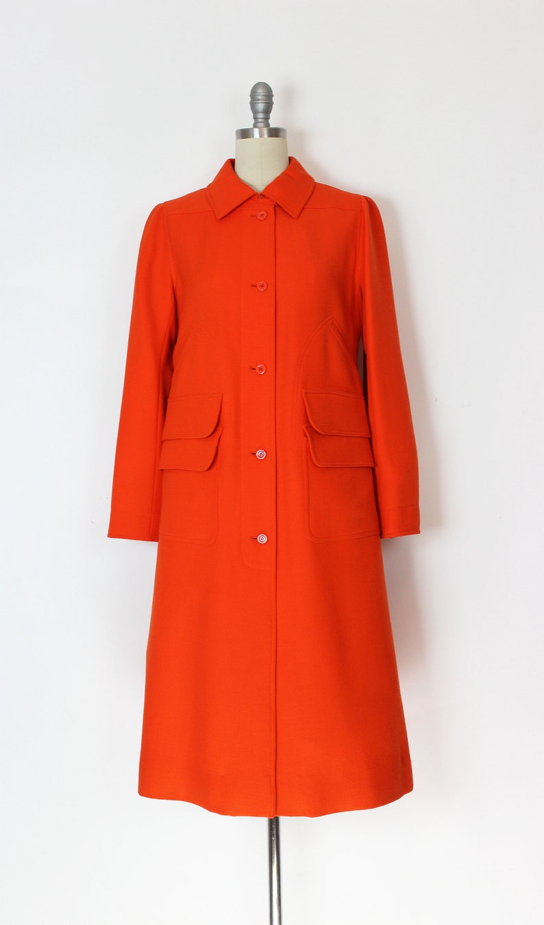May include: A long, bright orange coat with a button-up front and two large patch pockets on the front. The coat has a collar and long sleeves.