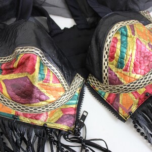 May include: Black bra with colorful, abstract patterned cups. The cups feature a mix of red, yellow, green, and purple hues, outlined with a cream-colored trim. Black fringe and beaded accents hang from the bottom.