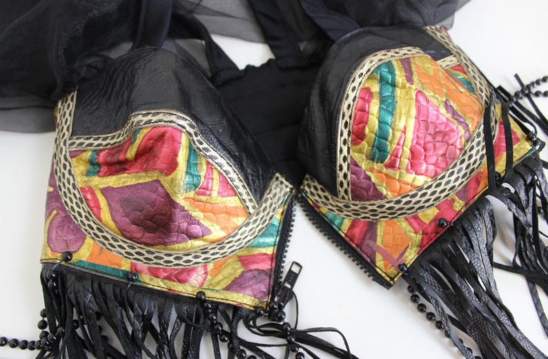 May include: Black bra with colorful, abstract patterned cups. The cups feature a mix of red, yellow, green, and purple hues, outlined with a cream-colored trim. Black fringe and beaded accents hang from the bottom.