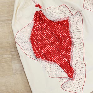 May include: Cream-colored skirt with red polka dot and decorative accents. The skirt features a red polka dot pattern on the front, with a decorative border and red trim. The skirt is made of a lightweight fabric.