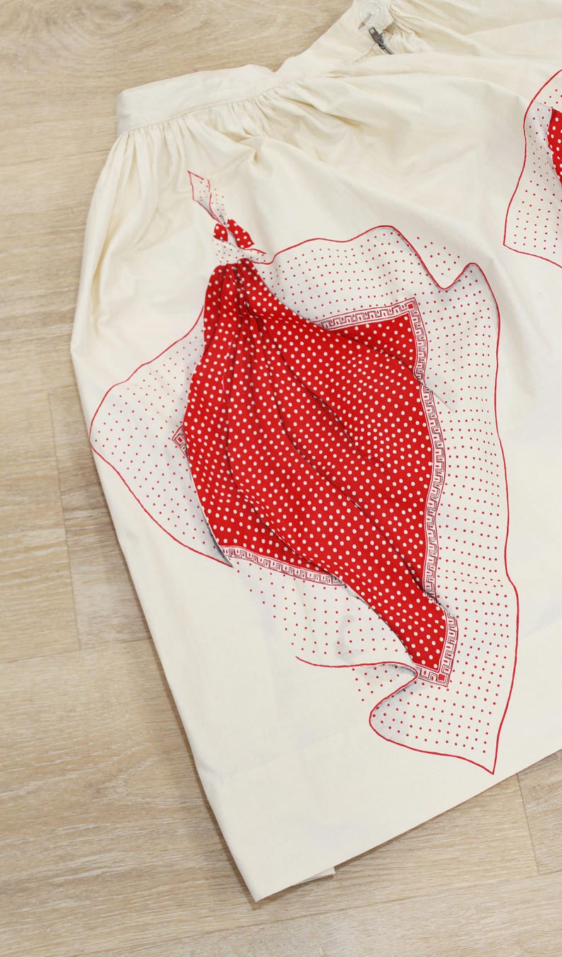 May include: Cream-colored skirt with red polka dot and decorative accents. The skirt features a red polka dot pattern on the front, with a decorative border and red trim. The skirt is made of a lightweight fabric.