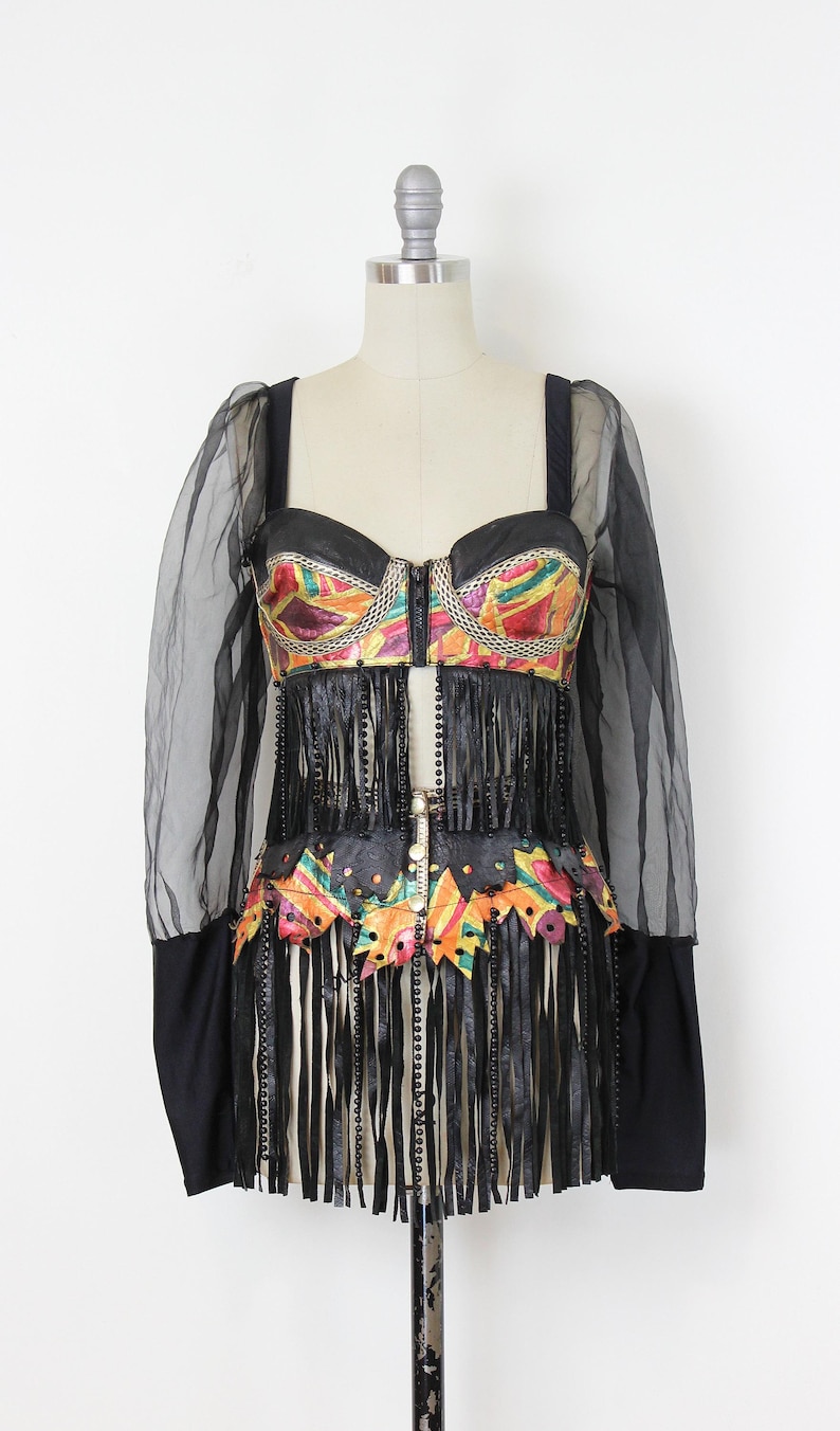 May include: A black and colorful vintage dress with a bustier top and fringe detailing. The top features a colorful geometric pattern and a zipper closure. The dress has long, sheer sleeves and is displayed on a mannequin.
