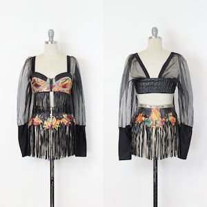May include: A two-piece black outfit on a mannequin. The top features a bustier with colorful embellishments and fringe, paired with sheer sleeves. The skirt has matching embellishments and fringe. The outfit is displayed against a white background.