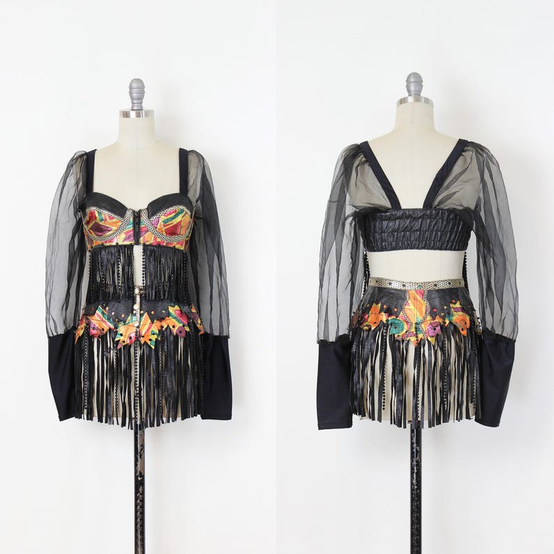 May include: A two-piece black outfit on a mannequin. The top features a bustier with colorful embellishments and fringe, paired with sheer sleeves. The skirt has matching embellishments and fringe. The outfit is displayed against a white background.