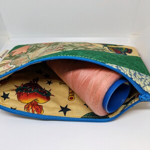 May include: A zippered pouch with a blue zipper and a green, floral, and beige patterned fabric exterior. The pouch is open and a spool of pink thread is inside.