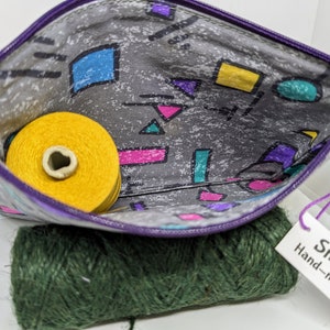 May include: A purple zippered pouch with a grey and white geometric print. The pouch is open and a yellow spool of thread is inside. The text on the tag reads "Small Purse Hand-made with Love".