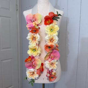 May include: A decorative stole featuring two vertical strands of artificial flowers in shades of pink, orange, yellow, and white. The floral arrangement includes orchids, poppies, and various blossoms accented with small beads and green leaves.