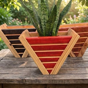 May include: Three wooden, triangular planters with a plant inside. The planters are made of natural wood and stained in various colors, including red, brown, and black. The planters are sitting on a wooden table.