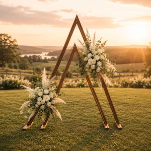 May include: A double triangular wooden wedding arch frame featuring two nested structures of varying heights. Each frame is decorated with arrangements of white roses, eucalyptus leaves, and tall pampas grass plumes attached to the wooden beams.