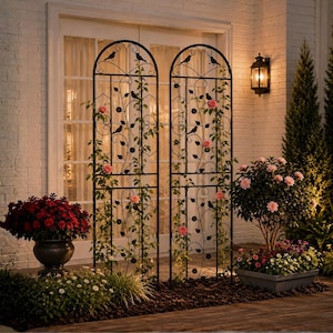 May include: Two tall, arched black metal garden trellises featuring decorative scrollwork, leaf motifs, and small bird silhouettes. The structures are adorned with climbing green vines and pink flowers, positioned in front of a white brick wall.