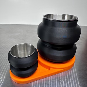 May include: A set of two black, rounded containers with metallic silver interiors, mounted on a bright orange, oval-shaped base. The larger container is positioned behind the smaller one, both featuring a stacked, tiered exterior design.
