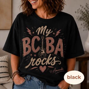 May include: A black short-sleeve t-shirt featuring a graphic design with the text "My BCBA rocks" in a distressed font. The design includes a central electric guitar illustration, lightning bolts, stars, and a heart. The name "Amanda" is printed below.