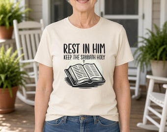 May include: A cream-colored short-sleeve t-shirt featuring a black graphic of an open book with a small cross above it. The text above the book reads "REST IN HIM" and the text below reads "KEEP THE SABBATH HOLY" in a bold, dark font.