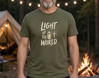 May include: An olive green short-sleeved t-shirt featuring the text "LIGHT OF THE WORLD" in a cream-colored font, accompanied by a graphic illustration of a lantern and a cross with radiating light lines centered on the chest.
