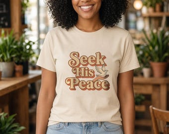 May include: A beige short-sleeve t-shirt featuring the text "Seek His Peace" in a brown and orange retro-style font, accompanied by a graphic illustration of a white dove resting on an open hand.