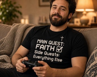 May include: A black short-sleeved t-shirt featuring white text that reads Main Quest: Faith with a checkmark symbol and a cross icon, followed by the text Side Quests: Everything Else, worn by a person holding a black game controller.