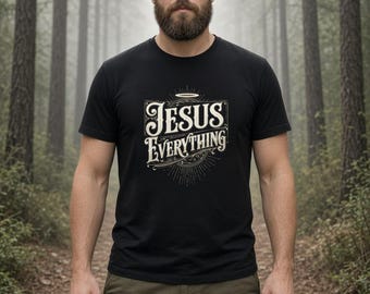 May include: A black short-sleeved t-shirt featuring a graphic print in white and gold tones. The design includes the text "JESUS" above the word "EVERYTHING" with a halo icon above the lettering and decorative line work surrounding the text.