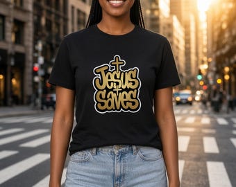 May include: A black short-sleeved t-shirt featuring a gold-colored graphic print with a white outline. The text reads "Jesus Saves" in a stylized font, with a small cross symbol positioned above the word Jesus and between the two words.
