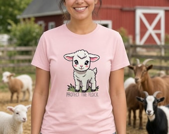 May include: A light pink short-sleeve t-shirt featuring a graphic illustration of a white lamb standing in green grass with the text "PROTECT THE FLOCK" printed in black lettering below the animal.