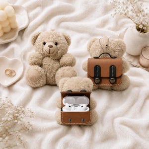 Peut inclure: Trois ours en peluche beiges, dont un avec un étui en cuir marron contenant des écouteurs blancs. Un autre ours a un petit cartable en cuir marron. Les ours sont sur une surface blanche texturée.