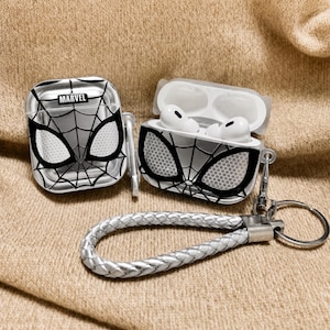 Peut inclure: Étui transparent pour AirPod avec un motif Spider-Man, présentant un motif de toile d'araignée et des détails d'yeux. L'étui est ouvert, révélant des écouteurs AirPod blancs. Une dragonne tressée argentée avec un fermoir en métal est attachée. Le mot "MARVEL" est imprimé.