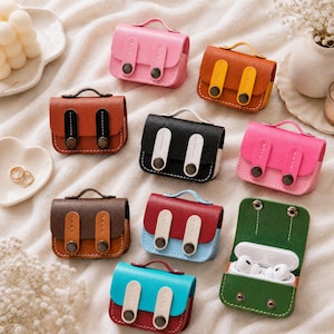 Peut inclure: Une collection de petits étuis en cuir colorés, conçus pour contenir des écouteurs sans fil. Les étuis sont de différentes couleurs, notamment rose, marron, noir et vert, et comportent une petite poignée et des fermoirs décoratifs. Un étui est ouvert, révélant des écouteurs blancs.
