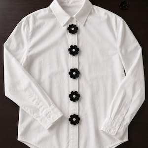 May include: A crisp white long-sleeve button-down shirt featuring a vertical row of five black crocheted flower embellishments centred along the placket. Each flower is accented with a small white button in the centre. Five additional loose black flowers are placed nearby.