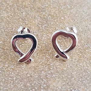 May include: A pair of silver-colored stud earrings. Each earring features a heart-shaped design with a ribbon-like detail at the bottom. The earrings are displayed on a neutral, textured surface.