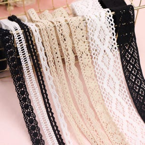 May include: A collection of lace trim ribbons in black, white, and beige. The assortment features various intricate woven patterns, including floral, geometric, and scalloped edges, displayed in parallel strips with diverse widths and textures.