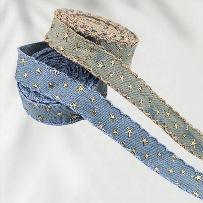 May include: Two spools of decorative ribbon. One is blue with gold star accents and a scalloped edge. The other is a muted green with gold star accents and a scalloped edge. The ribbon is made of a denim-like material.